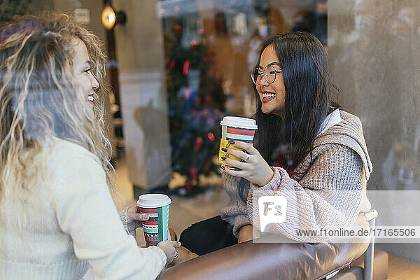 Young female friends having coffee while sitting in cafe seen through window glass