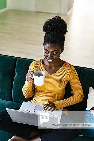 Smiling Afro woman using laptop while holding coffee cup in living room at home
