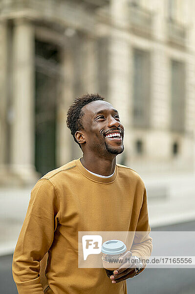 Smiling young man with disposable cup looking away while standing in city