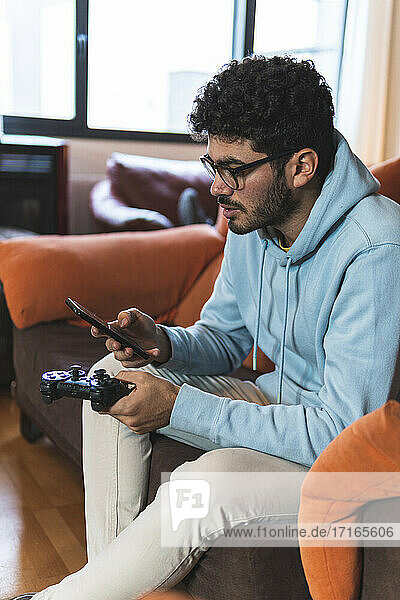 Junger Mann  der ein Smartphone mit einem Spiel-Controller benutzt  während er auf dem Sofa im Wohnzimmer sitzt