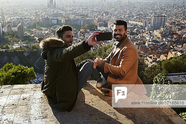 Mann  der ein Selfie mit seinem Freund macht  während er auf dem Aussichtspunkt vor der Stadtkulisse sitzt  Bunkers del Carmel  Barcelona  Spanien