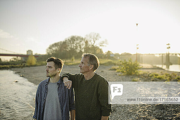 Father and son looking away while standing on riverbank at evening