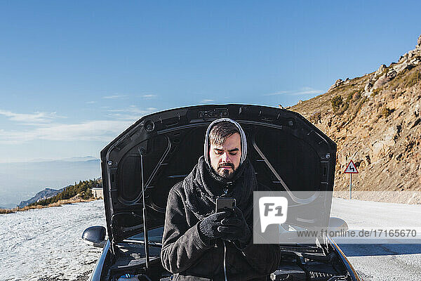 Man in warm clothing using mobile phone on snow covered land against vehicle hood