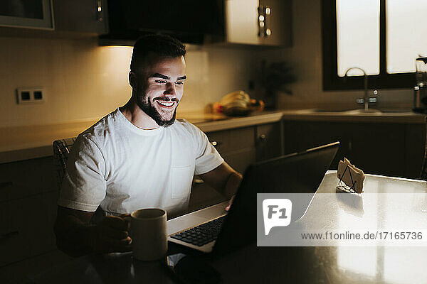 Bärtiger männlicher Unternehmer mit Kaffeetasse bei der Arbeit am Laptop am Esstisch in der Küche