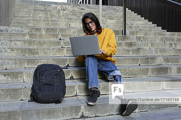 Smiling man with leg prosthesis using laptop while sitting on steps