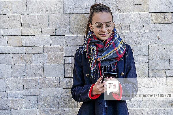 Teenage girl wearing warm clothing using mobile phone while standing against brick wall at home