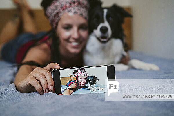Frau nimmt Selfie durch Handy  während auf dem Bett liegend mit Hund zu Hause