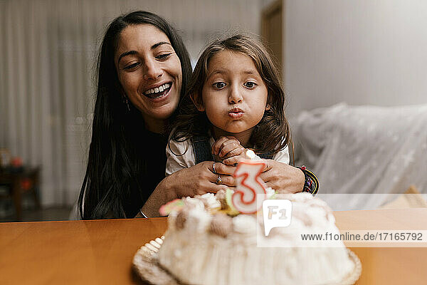 Mädchen bläst eine Kerze auf einem Geburtstagskuchen aus  während sie mit ihrer Mutter zu Hause sitzt