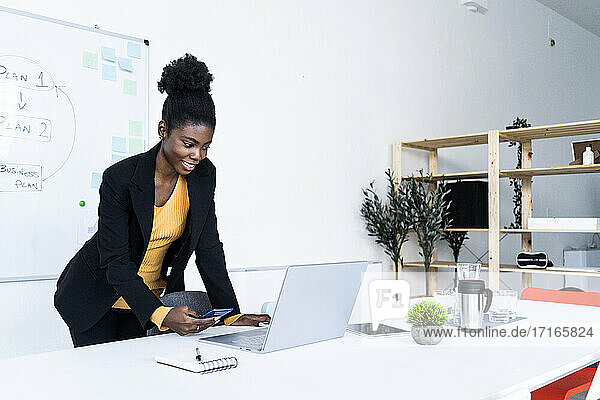Smiling young Afro businesswoman using smart phone while looking at laptop in office