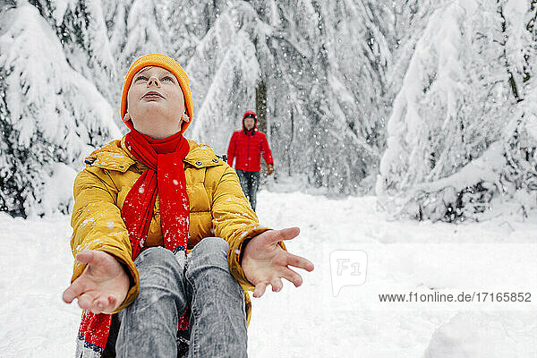 Boy looking up while sitting with man in background at forest during snowing