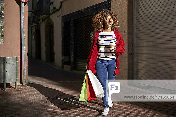 Smiling woman with coffee cup and shopping bag looking away while walking on footpath