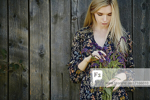 Beautiful blond woman holding bunch of purple flowers while standing against old wooden wall