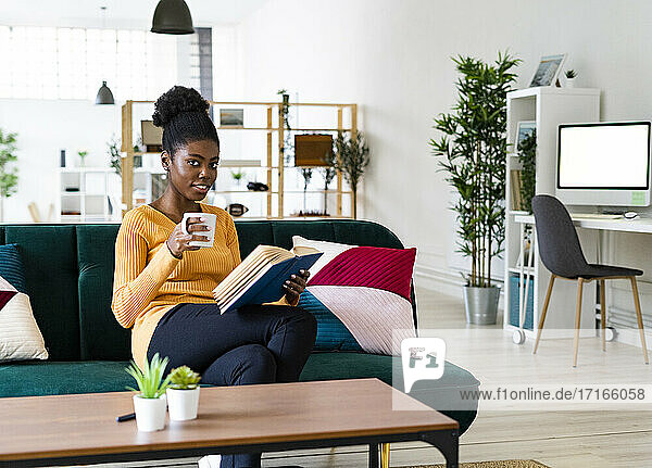 Young Afro woman reading book while sitting with coffee cup on sofa at home