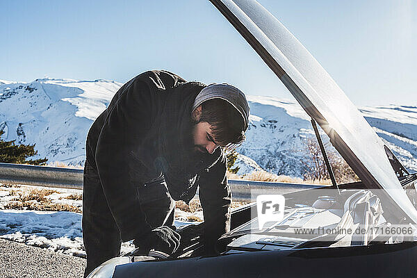 Man repairing car on snow covered land against sky during winter