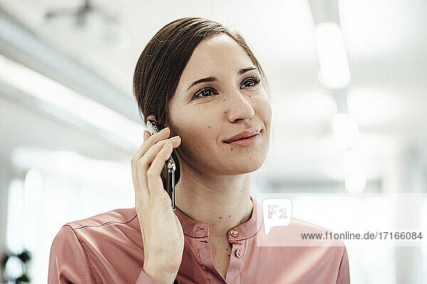 Lächelnde Geschäftsfrau  die wegschaut  während sie mit einem Smartphone im Büro spricht