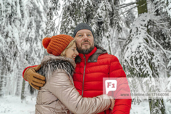 Smiling woman embracing man while standing in forest during winter