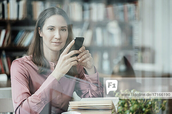 Female entrepreneur with book holding mobile phone looking through glass in cafe