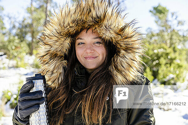 Lächelnde Frau mit Pelzkapuzen-Wintermantel  die eine Flasche hält  während sie im Wald steht