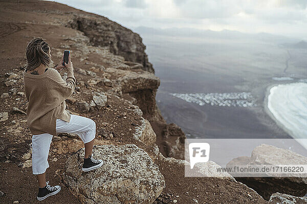Weibliche Touristin  die ein Foto mit ihrem Handy macht  während sie auf einem Berg am Strand von Famara steht  Lanzarote  Spanien