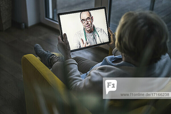 Ältere Frau führt ein Videogespräch mit einem männlichen Arzt über ein digitales Tablet im Wohnzimmer