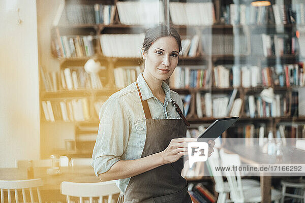Female owner holding digital tablet seen through window at coffee shop