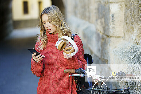 Blond woman with headphones using mobile phone while standing by bicycle