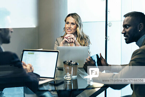 Smiling businesswoman with hand on chin listening to businessman while sitting in meeting at office