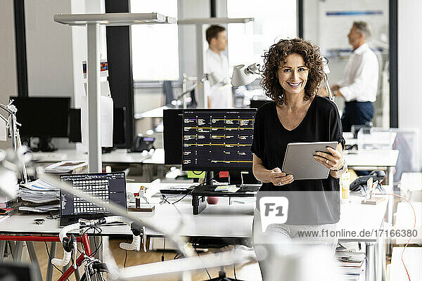 Lächelnde Geschäftsfrau  die ein digitales Tablet hält  während sie mit einem Kollegen im Hintergrund in einem Großraumbüro steht