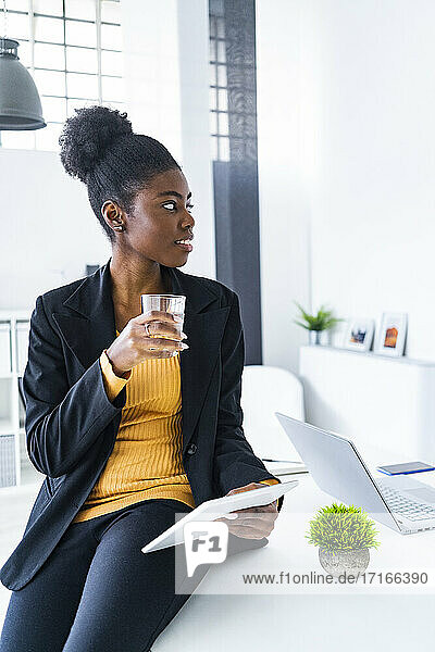 Young female Afro professional looking away holding glass of water and digital tablet while sitting on desk