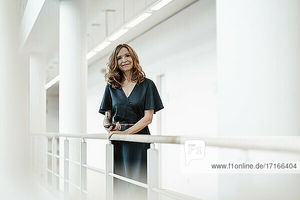 Lächelnde Geschäftsfrau mit Smartphone am Geländer lehnend im Korridor