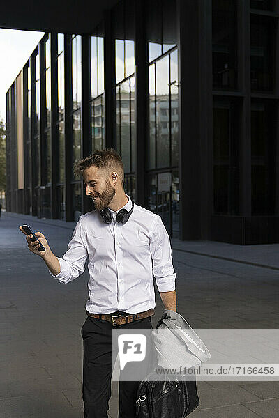 Businessman holding briefcase while using mobile phone standing on footpath