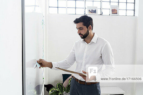 Young businessman with clipboard sticking adhesive note on whiteboard at work place