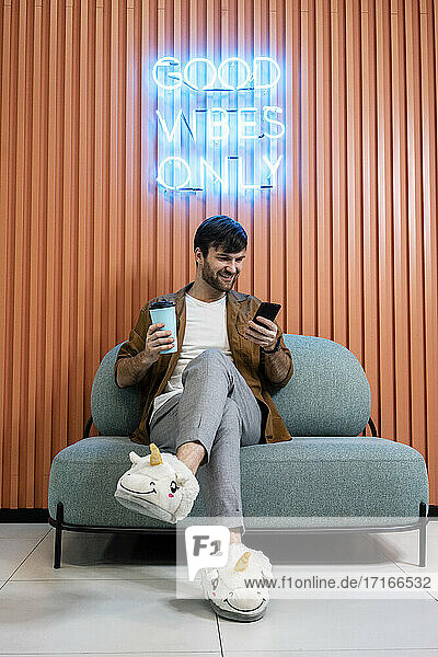 Smiling creative businessman with coffee cup in unicorn slippers using smart phone against wall in office