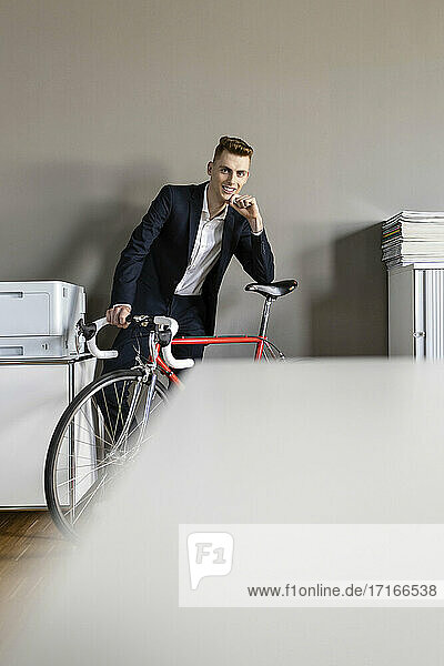 Confident businessman with hand on chin leaning on bicycle while standing at office