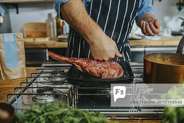 Chef checking raw tomahawk steak in frying pan while standing in kitchen