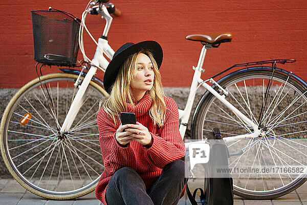 Woman with mobile phone looking away while sitting against bicycle