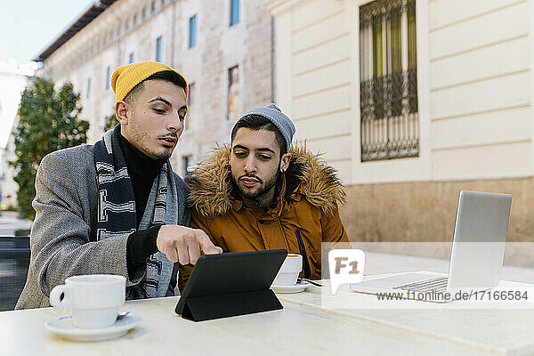 Junges schwules Paar  das in einem Straßencafé sitzt und ein digitales Tablet benutzt