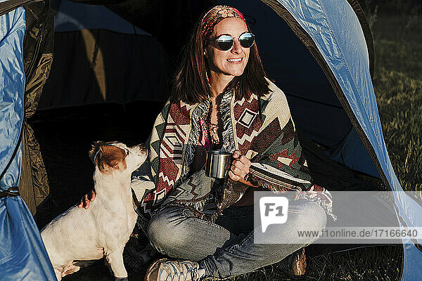 Glückliche Frau mit Sonnenbrille  die eine Tasse hält  während sie mit ihrem Hund im Zelt sitzt