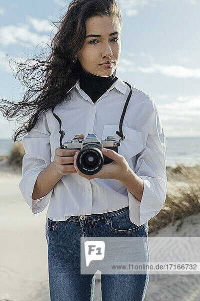 Beautiful young woman holding camera while standing at beach against sky