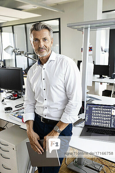 Selbstbewusster Geschäftsmann mit digitalem Tablet  der am Schreibtisch im Büro steht und starrt