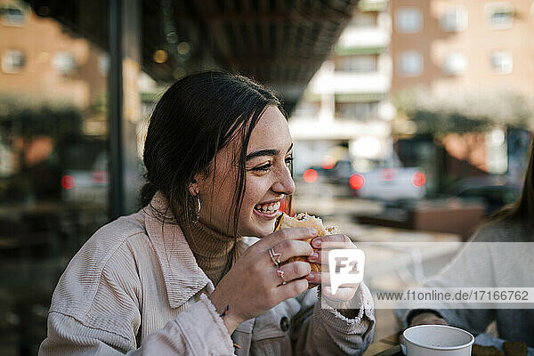 Close-up of cheerful teenage girl eating burger with friend at sidewalk cafe