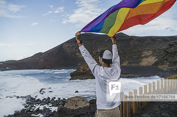 Junger männlicher Tourist  der die Regenbogenfahne schwenkt  während er am Aussichtspunkt El golfo  Lanzarote  Spanien steht