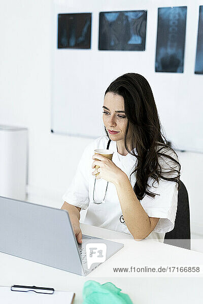 Young female doctor having coffee while using laptop in clinic