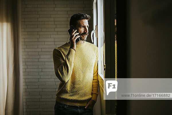 Mid adult man looking through window while talking on mobile phone at home