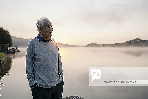 Senior man looking at view while standing against lake during sunrise