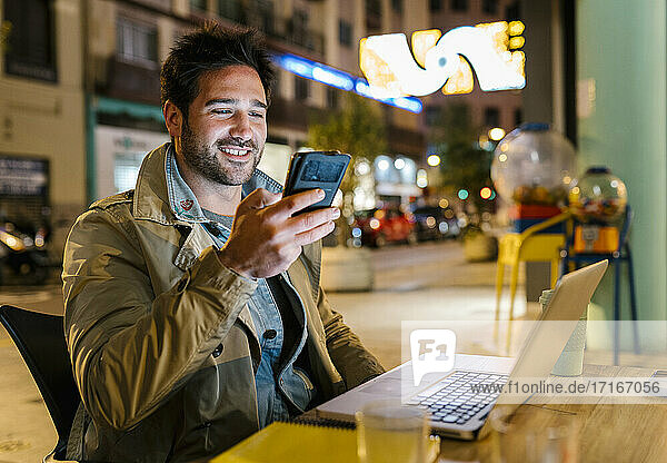 Smiling man with mobile phone using laptop while sitting at sidewalk cafe