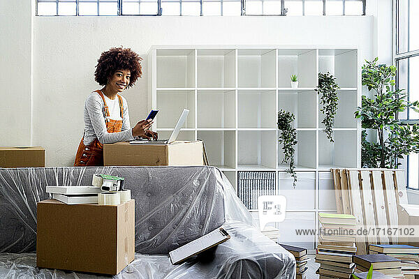 Smiling young woman standing with smart phone and laptop while moving into new home
