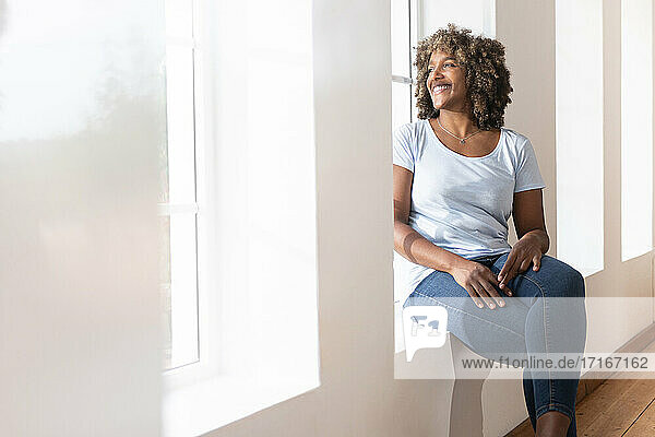 Mittlere erwachsene Frau schaut weg  während sie zu Hause auf der Fensterbank sitzt