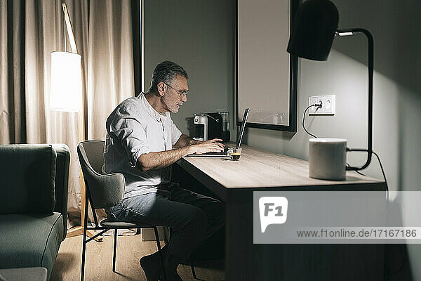 Mature man sitting at desk with coffee while using laptop in hotel room