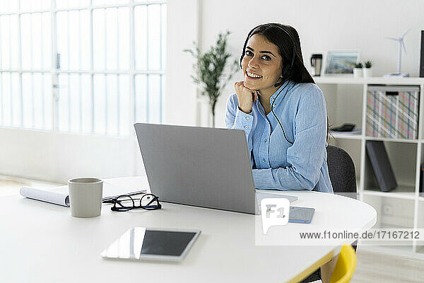Businesswoman wearing headset smiling while sitting with laptop at office